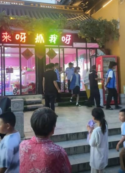 图片拍摄于一家夹娃娃机店门口，门头写着“来呀抓我呀”，门口正有警察带人走，形成了现实与招牌文字的幽默巧合。店招原意招来顾客“抓娃娃”，但此刻仿佛在回应警察执法的场景，充满双关和戏剧感。有不少路人在台阶下看热闹，整体氛围既有紧张又带点荒诞、出其不意。