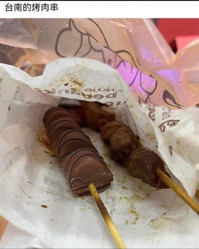 这是一张玩食物反差梗的搞笑图，标题标注为「台南的烤肉串」，但打开包装后，却呈现出强烈的视觉反差：一根是正常的烤肉串，另一根却是用巧克力块串成的「仿烤肉串」，巧克力被做成类似烤肉串的形态，和旁边的真烤肉串形成趣味对比，制造出意外的笑点。
无谐音梗或双关，纯靠食物的反差感带来趣味效果。