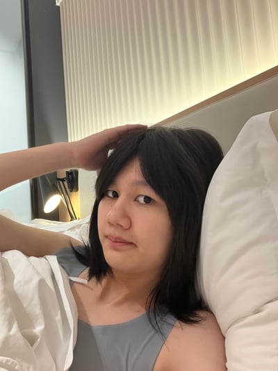 这是一张真人实拍的竖版照片，拍摄场景为酒店卧室。画面中一名留黑色中短发的女性靠在床头的白色枕头上，身穿灰色吊带内搭，身上盖着部分白色被褥，一只手抬起放在自己头顶，表情放松看向镜头。背景可见竖条纹装饰的床头墙板，还有一盏亮着的黑色壁挂台灯，整体氛围慵懒随意。