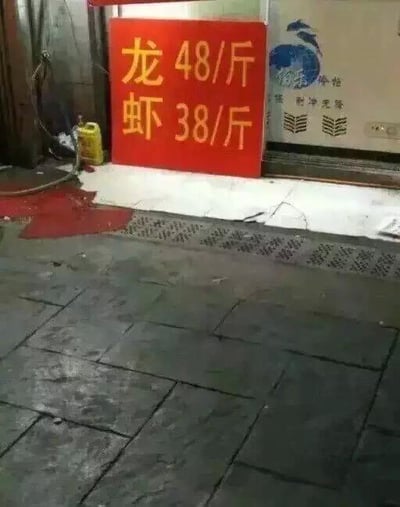 这是一张在街边餐饮店铺门口拍摄的实拍图，画面主体是一个红色的价格立牌，立牌上用醒目的黄色字体分两行标注“龙 48/斤”“虾 38/斤”，实际是商家将“龙虾”二字拆分成两行排版，制造出“龙”与“虾”分开定价的视觉错觉，本质是龙虾的不同规格或不同做法的两种售价，用这种趣味化的排版方式吸引路人注意力，属于一种接地气的餐饮营销小创意，无其他特殊谐音或暗示内容。
