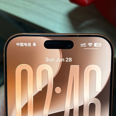 这是一张带有灵动岛设计的金色款苹果手机的锁屏界面实拍图，画面左下角可看到握持手机的手指边缘，背景模糊。锁屏界面顶部左侧显示运营商为中国电信、静音提醒标识，右侧为信号、WiFi标识以及43%的电量显示；中间位置标注日期为“Sun Jun 28”，下方是超大字号的02:46时间显示，锁屏壁纸为同色系暖棕色渐变的抽象几何图案。该内容利用英文日期缩写“Sun Jun”（六月周日的缩写）与中文人名“孙俊”拼音发音一致的特点构成谐音趣味点。