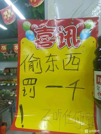 这是一张拍摄于超市内的图片，主体是一张原本设计为喜庆风格的“喜讯”告示模板，却被用黑色粗笔手写上了“偷东西 罚一千 不听任何理由”的防盗警示内容，顶部的红色“喜讯”字样和下方严肃的警示语形成强烈反差。背景中可以看到超市的货架，上面摆放着各类商品，右侧边缘有一瓶海天金标生抽，整体场景是普通的超市内景，这张另类的防盗告示显得格外醒目又带有讽刺感。