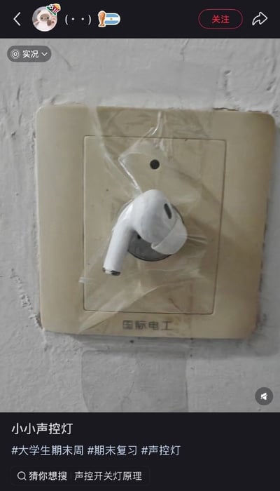 这是一张社交平台分享截图，画面主体是安装在灰白色水泥墙面上的米黄色“国际电工”牌声控灯开关面板，有人用透明胶带将一只白色AirPods耳机粘贴在声控灯的拾音孔位置，利用耳机的拾音功能放大轻微声响来触发声控灯点亮。截图下方配文“小小声控灯”，附带#大学生期末周、#期末复习、#声控灯的话题标签，底部还有搜索推荐“声控开关灯原理”。该操作是大学生期末复习阶段的趣味小妙招，避免夜晚出声喊亮声控灯打扰到其他室友。