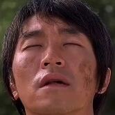 图片为周星驰的面部特写，他双眼紧闭，眉头微蹙，脸上带有明显的污渍，表情显得有些疲惫或痛苦。背景为模糊的绿色，可能是户外场景。