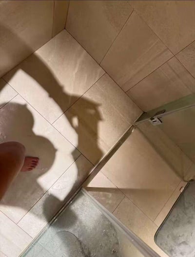 这是一张在浴室中拍摄的趣味照片，画面左下角露出拍摄者的小腿与赤脚，阳光在米白色瓷砖地面投射出两处影子，其中一处因光影错位巧合形成了类似猫咪的轮廓，和拍摄者的影子相邻，制造出仿佛有猫咪在旁的视觉错觉，让普通的日常场景充满了意外的趣味感。