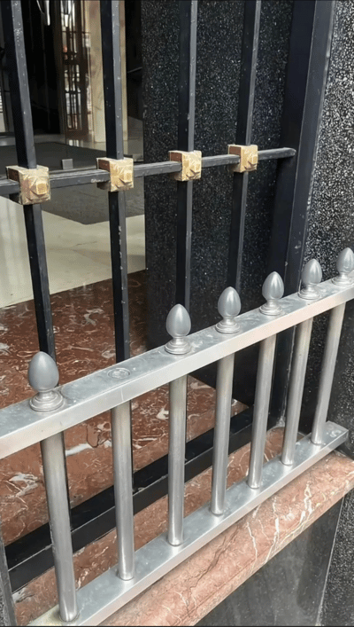 这是一张建筑细节特写照片，展示了一扇金属大门的局部结构。画面中可见上下两部分栏杆：上部为黑色竖栏，通过三个金色方形紧固件横向连接；下部为银色竖栏，顶端呈尖锥形，底部固定在带有大理石纹理的地面上。背景中能看到深色的颗粒状墙面和门内的部分空间，整体呈现出现代建筑的质感与工艺细节。