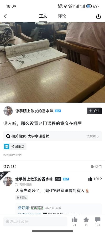 这张截图是社交平台的发帖页面，上方是拍摄者在大学教室拍摄的场景：浅棕色课桌上面摊放着一本翻开的满是文字的书本，书本上放着一副眼镜，周围能看到其他学生的座位和身影，能看出是大学课堂的环境；下方是发帖内容，用户头像是动漫人物，昵称为“像手腕上散发的香水味”，发帖内容为“没人听，那么设置这门课程的意义在哪里”，内容关联标签为校园生活，相关搜索词条是“大学水课现状”；该用户后续发布置顶评论玩谐音梗，内容为“大家先别吵了，我刚在教室里看到有人🦌”，用“鹿”的谐音指代“录（音/录屏）”，暗示课堂上有人正在记录相关发言，提醒大家停止讨论，该评论获得1012个点赞，评论区共有184条评论。