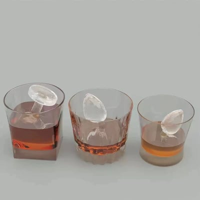 展示了三个装有琥珀色液体的玻璃杯，每个杯子中都有一个独特形状的冰块。杯子从左到右依次排列，背景为简约的灰色，突出展示了饮品和冰块的质感。