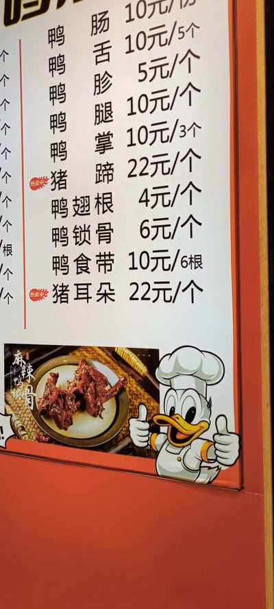 图片展示了一个橙色边框的立式小吃摊价目表，顶部有部分店铺名称文字（不完整）。主体为白色背景黑色文字的价格列表，包含多种鸭货及猪副产品：鸭肠10元/份、鸭舌10元/5个、鸭胗5元/个、鸭腿10元/个、鸭掌10元/3个、猪蹄22元/个（标注“爆款”）、鸭翅根4元/个、鸭锁骨6元/个、鸭食带10元/6根、猪耳朵22元/个（标注“爆款”）。价目表下方左侧配有一盘麻辣鸭货的实物图片，标注“麻辣鸭哥”；右侧是一个戴着厨师帽、穿着白色制服的卡通鸭子形象，正咧嘴大笑并竖起双拇指点赞，整体设计活泼亲切。