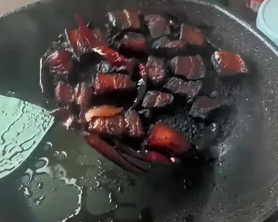 这是一张厨房烹饪翻车的实拍图，铁锅里的红烧肉完全炒糊碳化，大部分肉块变成焦黑色，仅少量残留原本的肉色，锅面和肉块上附着焦糊痕迹，一旁的锅铲沾有油迹，显然是烹饪时疏忽火候导致的失败作品，充满无奈又搞笑的氛围。