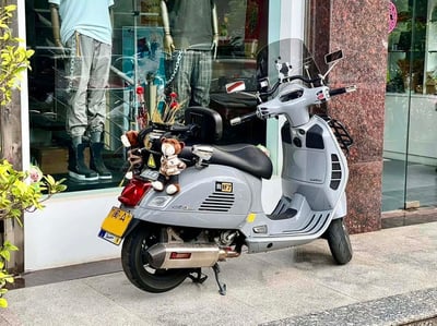 图片中是一辆停放在商店橱窗前的灰色小踏板摩托车，车身经过个性改装，车头和车尾放置了可爱的小熊玩偶。摩托车侧面贴有“我好了”字样的贴纸，车牌号为“闽A”（推测为福州车牌）。背景中可以看到商店橱窗内的人体模特，穿着休闲服装，橱窗玻璃反射出周围环境。摩托车旁有绿色植物点缀，整体画面充满生活化气息，展现了车主的个性品味。
