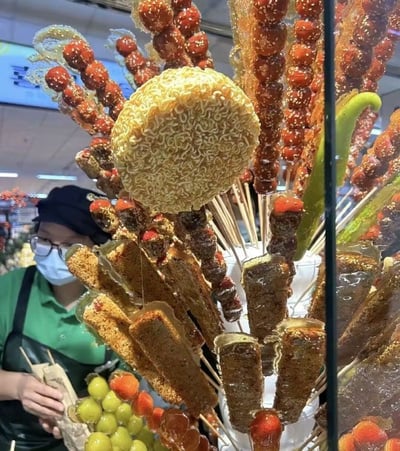 这张照片展示了一个创意冰糖葫芦摊位，传统的山楂冰糖葫芦与各种新奇食材混搭在一起。最引人注目的是一串圆形方便面饼被制成冰糖葫芦，周围环绕着传统的红色山楂串、绿色黄瓜条和其他水果串。所有食材都裹着晶莹剔透的糖衣并撒有芝麻，在灯光下显得格外诱人。背景中可以看到一位穿着绿色上衣、戴着黑色围裙和口罩的摊主，正在整理这些特色小吃。整个场景呈现出中国街头小吃的创新与传统结合的特点。
