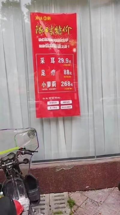 图片展示了一家店铺的红色广告招牌，上面用黄色和白色字体写着各种服务项目和价格。招牌顶部写着'耳沐①新 限时特价'，中间列出了三项服务：采耳29.9元（30分钟）、足疗88元（60分钟）和小萝莉268元（90分钟）。招牌底部还有店铺地址电话和'专业服务、品质保障、贴心服务、舒适环境'等宣传语。招牌被贴在玻璃门上，下方可以看到一辆摩托车的部分车身。