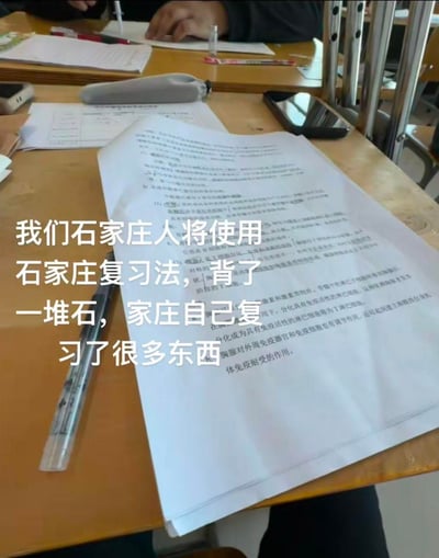 图片展示了一张摆满复习资料的课桌场景，桌上有写满字的试卷、钢笔、手机等学习用品，背景中可见同学正在低头书写。画面中央覆盖着醒目的白色文字：“我们石家庄人将使用石家庄复习法，背了一堆石，家庄自己复习了很多东西”。

该图片运用了谐音梗，将“石家庄”拆分为“石”和“家庄”，调侃式地创造出“背了一堆石（实际没记住），家庄（谐音‘自己’）复习了很多东西”的幽默效果，反映学生群体对复习效率的自嘲。