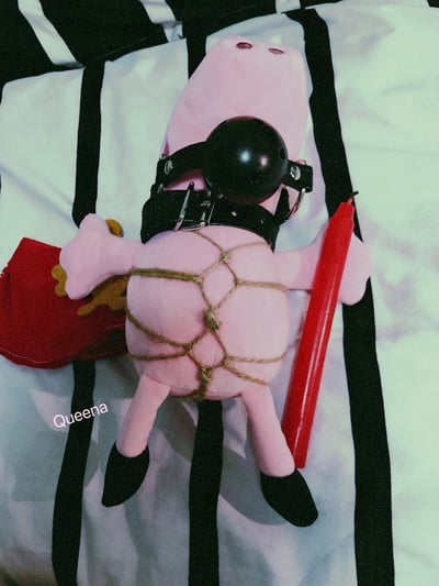 这是一张静态实拍图，主体是经典儿童动画《小猪佩奇》的同款粉色毛绒玩偶。玩偶被麻绳以绳艺的方式捆绑住躯干，脖子佩戴着带金属扣的黑色皮质项圈，头部戴着黑色的口塞类束缚道具，面部被遮挡。玩偶的一只手握着一支红色长蜡烛，另一只手拿着一个带有黄色装饰的红色小包，平躺于黑白条纹的床品之上。图片左下角带有白色的“Queena”水印，整体是对儿童向玩偶进行的另类恶搞创作，风格猎奇有趣。