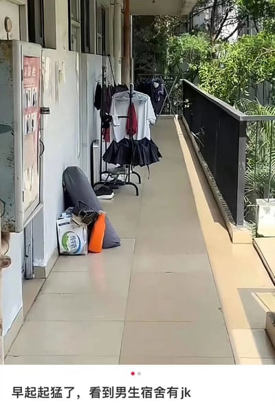 这张图片展示了一个男生宿舍的走廊场景。走廊地面铺着浅色瓷砖，左侧墙边放着一个晾衣架，上面挂着白色上衣和黑色百褶裙（类似JK制服），旁边还有灰色的包、鞋子、印有“纯牛奶”字样的盒子和橙色容器等日常物品。走廊右侧是黑色栏杆，外面能看到绿色植物。图片底部配有文字“早起起猛了，看到男生宿舍有jk”，以幽默的方式表达了早起时看到男生宿舍出现JK制服的惊讶与误会，实际上可能只是室友的衣物。