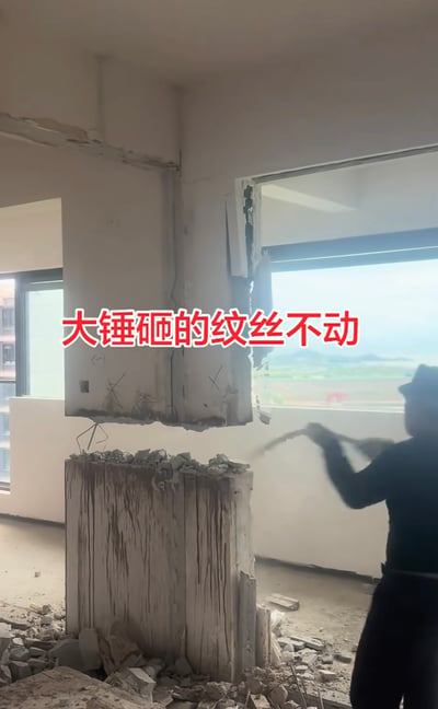 图片展示了一处装修拆除现场：一名身穿深色衣物的工人正背对镜头，手持大锤奋力砸向房间角落的墙体。墙体底部已被砸出缺口，地面散落大量混凝土碎块，但上部墙体依然坚挺。画面中央有红色文字覆盖“大锤砸的纹丝不动”，直观呈现拆除工作遇阻的场景。结合原始标题推测，该墙体可能为承重结构，因此异常坚固导致难以拆除。