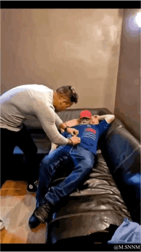 这是一段搞笑的动态GIF，画面中一名穿浅灰色上衣的男子站在黑色皮沙发旁，伸手去扯躺在沙发上的男子的裤子；躺卧的男子身着蓝色上衣与牛仔裤，头戴红色帽子，双手枕在头后，看起来放松又带点戏谑，似乎在配合这场朋友间的恶作剧，整体氛围轻松欢乐，充满整活趣味。