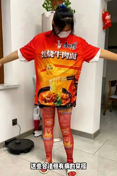 图片中展示一位女性身着极具创意的“食品包装风”穿搭：上身是红色康师傅红烧牛肉面主题T恤，完整复刻了泡面包装设计，印有“康师傅红烧牛肉面”大字及面条、牛肉、蔬菜的诱人餐食图像，辅以“美味升级 经典口味”标语；下身搭配红色双汇火腿 leggings，布满品牌标识及产品信息。人物双臂向两侧平伸，站立于室内，背景有银色冰箱、绿植、吸尘器及红色卡通挂饰，面部佩戴白色口罩。底部文字“这些合法但有病的穿搭”以幽默口吻点出此造型虽合规却视觉荒诞的特点。整体通过日常食品包装与服装的结合，营造出强烈的喜剧效果和话题性。