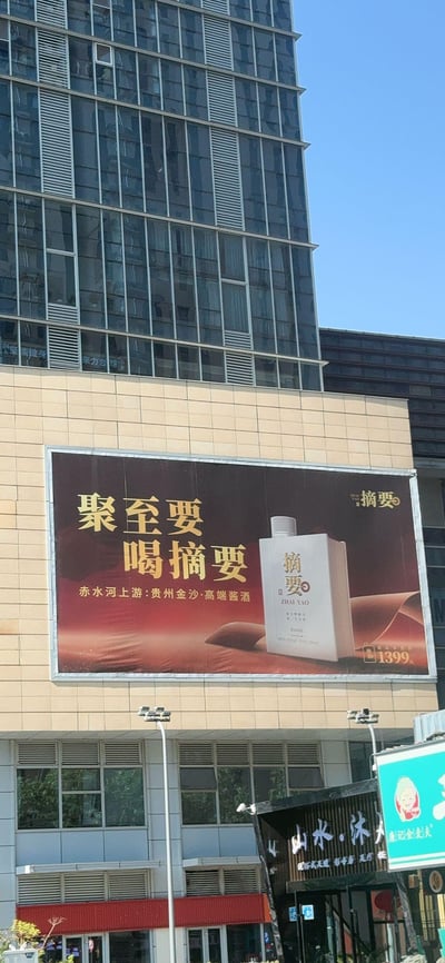 这是一张户外实拍的楼体广告图，画面主体是贵州金沙摘要酒的宣传广告牌，安装在现代玻璃幕墙建筑的低层外墙上。广告牌以红棕渐变底色搭配金色、白色文字，突出宣传语“聚至要 喝摘要”，同时标注了“赤水河上游:贵州金沙·高端酱酒”，并展示了简洁的白色酒瓶样式，标明建议零售价1399元。广告牌下方能看到临街的商铺，背景是晴朗的蓝天，整体呈现出城市户外商业宣传的正式场景，没有双关、谐音梗或暗示内容。