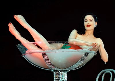 著名 burlesque 表演者蒂塔·万提斯（Dita Von Teese）的经典舞台造型：她身着白色羽毛装饰，斜倚在巨型马提尼酒杯中，杯中盛满白色液体并漂浮着一颗草莓。她的双腿优雅抬起，红色脚趾甲与鲜艳红唇形成呼应，黑色复古波浪卷发搭配水钻发饰，背景为纯黑色，凸显出她标志性的复古性感风格。