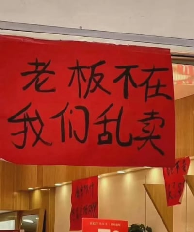 这是一张拍摄于室内卖场或店铺的照片，画面最上方悬挂着一张大红色的手写横幅，用黑色粗体字写着“老板不在 我们乱卖”，字体书写风格随意、接地气，充满趣味。在横幅下方还能看到几张尺寸较小的红色促销标语，部分文字被遮挡无法完全辨认。整体场景的装修风格偏简约，墙面有木质装饰，灯光柔和，营造出一种日常店铺的氛围。这条标语用夸张搞笑的话术，暗示店内正在进行不受约束的低价促销，以此吸引顾客注意，属于非常接地气的创意营销方式。