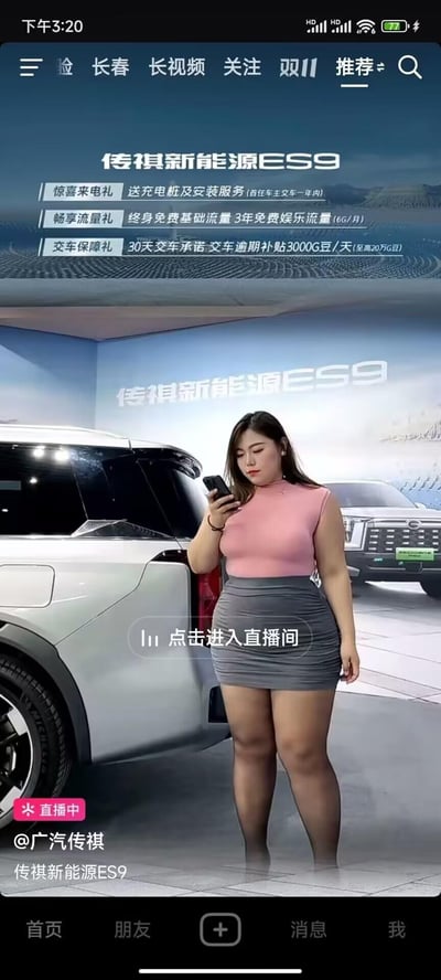 这是一张抖音平台的汽车品牌直播画面截图，展示了广汽传祺新能源ES9的线上营销场景。画面中一位微胖的女性主播身着粉色无袖上衣与灰色包臀裙，正低头专注查看手中的手机；背景是传祺新能源ES9的展车与品牌宣传背景墙，画面上方标注了该车型的三项购车福利：首任车主一年内赠送充电桩及安装服务、终身免费基础流量加3年每月6G娱乐流量、30天交车承诺且逾期每天补贴3000G豆（至高20万G豆）。画面下方带有“点击进入直播间”的引导按钮，左下角标注“直播中”与“@广汽传祺”的官方账号信息，正值双11营销节点，整体是品牌的新能源汽车直播带货场景。