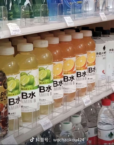 这是一张实拍的超市饮料货架照片，货架中层整齐摆放着多瓶外星人品牌的B族维生素饮料，分为青提口味（浅黄色液体）和橙子口味（橙黄色液体），旁边还陈列着外星人电解质水和其他瓶装饮用水。这款维生素饮料的瓶身标签存在排版设计问题，中文部分将"B水"放大放在标签上方，原本完整的英文单词"VITAMIN B"前半部分被遮挡，只露出后半段"AMIN B"，形成了滑稽怪异的文字排列效果，图片底部带有抖音号水印。