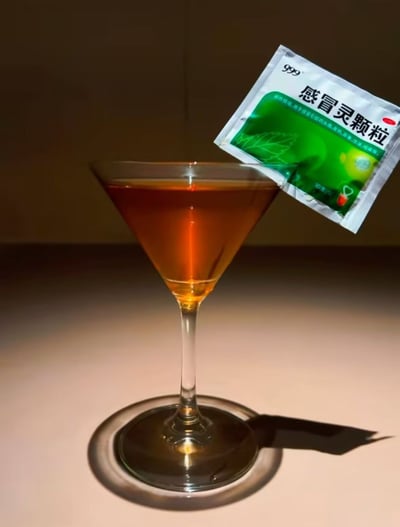 这是一张创意摆拍的静态照片，画面主体是一只透明的高脚鸡尾酒杯，杯中盛放着棕黄色的液体，是冲调好的999感冒灵颗粒，杯沿处搭着一包未拆封的999感冒灵冲剂。整体打光柔和，在浅色台面上投射出清晰的阴影，将普通的感冒药品用调酒的形式进行呈现，形成了生活化物品与精致调酒形式的反差感，充满趣味搞怪的网感风格。