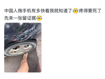 整张图为图文混排的社交媒体动态，上方配文为“中国人掏手机有多快看我就知道了🥹疼得要死了 先来一张留证据🥹”，下方实拍图显示拍摄者的脚被压在汽车轮胎下方，同时拍摄到了车身的剐蹭痕迹，以自嘲的方式调侃大家遇到意外事故时普遍会第一时间掏出手机拍摄留存证据的行为，充满趣味。