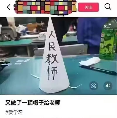 这是一张静态单图，拍摄于办公或学习的桌面场景，桌面上摆放着一个用白纸折成的尖顶锥形帽子，帽子正面用黑色手写字体写着“人民教师”四个大字。图片下方配有文字“又做了一顶帽子给老师”，并带有#爱学习的话题标签。这个尖顶帽的造型类似过去用来调侃表现不佳学生的“惩罚帽”，将其与“人民教师”的身份结合，形成了反差感，制造出搞笑的调侃效果，属于无恶意的趣味创作，用来玩师生日常的梗。