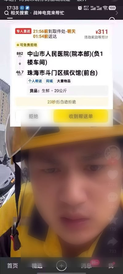 这是一张外卖骑手的直播画面截图，画面下半部分是身着黄色工作服、头戴黄色头盔的外卖员，面露凝重困惑的表情正对镜头，上半部分叠加了同城配送平台的派单弹窗：取件地址为距骑手882米的中山市人民医院院本部负一楼车间，收件地址为46.7公里外的珠海市斗门区殡仪馆前台，配送货品是20公斤生鲜，配送费311元，要求当晚21:56前取件、次日凌晨1:54前送达，订单标注为个人帮送、大重物品，支持免费拒绝，弹窗显示23秒后将自动拒单，下方配有「拒绝」和「收到帮送单」两个操作选项，画面底部是短视频平台的导航栏，包含首页、精选、发布、消息、我的入口。