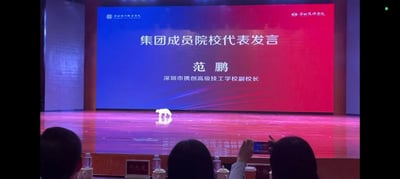 这是一个室内院校活动的会场舞台场景，舞台背景的蓝红渐变大屏上清晰显示着“集团成员院校代表发言 范鹏 深圳市携创高级技工学校副校长”的字样，大屏左上角带有“深圳城市职业学院”的标识，右上角则是“深圳技师学院”的标识。光亮的木质舞台地板反射着灯光，一只白色猫咪正安静地站在舞台中央，打破了原本严肃的发言场合氛围。台下能看到几位观众的背影，其中一位观众正举着手机拍摄这个意外的画面，前排的座位上摆放着名牌、水杯和矿泉水，其中一个名牌上写着“邓元龙”，整体场景在正式中透着意外的趣味感。