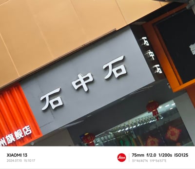 这是一张由小米13手机拍摄的线下店铺招牌实拍图，画面主体为灰色背景上的白色立体字“石中石”，左侧橙色区域可见“州旗舰店”字样，右侧玻璃门反射出“石中石”字样，店内悬挂有红色灯笼装饰。图片底部带有小米13、徕卡的标识及详细拍摄参数，包括拍摄时间2024年7月10日15:10:17，拍摄经纬度31°46'40"N 119°56'57"E，对应江苏常州地区。
