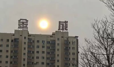 一张实拍城市建筑照片，展示了一栋多层居民楼的顶部景观。楼顶两侧分别矗立着金属材质的“金”和“成”立体大字，中间天空位置正有一轮明亮的太阳（疑似日出或日落），阳光透过略带灰蒙的天空形成明显光晕。画面右侧可见几棵无叶的枯枝树木，整体构图呈现出城市建筑与自然天象的偶然同框效果，氛围宁静而略带萧瑟。