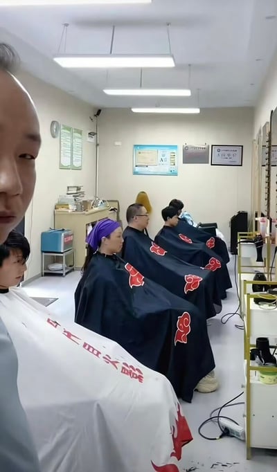 这张图片拍摄于一间类似教室或理发店的场所，几位理发顾客统一披着印有《火影忍者》中“晓组织”标志的披肩坐成一排，画面极具二次元反派团体的既视感，非常有趣好玩。场景中，披肩上的红云logo直接对应动漫里的经典反派组织，围观者很容易联想到“反派团建”等网络梗，既有cosplay趣味，也有社交玩梗属性。

双关/谐音梗说明：披着“晓组织”披风集体理发，搞笑还原动漫中经典排排坐画面，暗示大家也可能是“隐藏的反派”，具有很强的二次元自嘲意味。