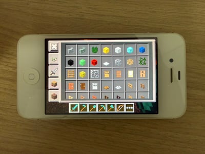 这张实拍照片呈现了一台带有实体Home键的老式苹果iPhone手机平放在浅棕色木纹桌面上，手机屏幕正显示着沙盒游戏《我的世界》的老版本移动端物品栏界面：界面左侧是物品分类按钮，中间网格区域整齐排列着各类像素风格的方块、门、栅栏等游戏道具，底部是玩家的快捷道具栏，摆放着剑、镐等工具，是很多《我的世界》老玩家十分熟悉的经典界面，极易引发怀旧情绪。