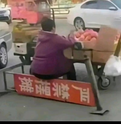 这是一张街头实拍的静态图，画面中一位穿着紫色连帽衫的摊贩，将写有'严禁摆摊'的红色警示标识牌当作座椅，坐在上面开展摆摊生意，标识牌的禁令内容和摊贩的行为形成强烈的反差，营造出荒诞又搞笑的效果，背景能看到停放的车辆和其他货物推车，是充满生活趣味的真实场景。