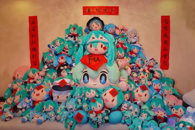 图片展示了大量初音未来主题的毛绒玩具被堆叠成金字塔形状，形成一个喜庆的新年装饰堆头。中央是一个大型初音未来毛绒玩具，胸前佩戴着红色