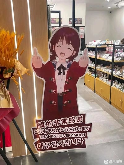 这是一张在二次元周边店铺内拍摄的照片，画面主体是一个经过二次创作的动漫人物立式人形牌，人物为《偶像大师 闪耀色彩》中的樋口圆香，她穿着酒红色牛角扣大衣搭配水手服内搭，双眼紧闭、嘴巴大张，做出类似双手比中指的动作，但下方的标牌用中文、日文、英文、韩文四种语言写着“真的非常感谢”“どうもありがとうございます”“THANK YOU VERY MUCH”“매우 감사합니다”，动作与文字形成强烈反差，制造出搞笑效果。店铺背景里可见货架上摆满了各类二次元周边商品，画面左侧还有装饰用的麦穗摆件。