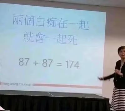 这是一张职场培训现场的实拍图，画面右侧是一位女性讲师，她手持话筒，左手伸出指向左侧的投影屏幕，正在进行讲解。投影屏幕上用繁体中文写着“兩個白痴在一起就會一起死”，下方搭配了数学算式“87 + 87 = 174”。这里运用了粤语谐音梗：数字“87”的粤语发音和“白痴”相近，“174”的粤语发音和“一起死”相近，用数学计算的形式包装粗俗玩笑，制造出荒诞搞笑的效果。