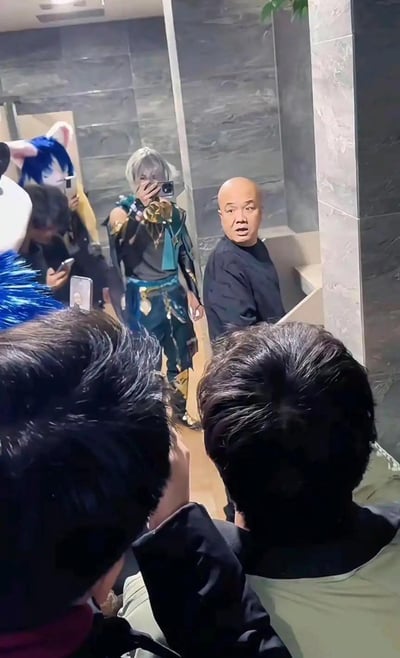 这是在公共卫生间镜子前的实拍场景：一群穿着二次元cos服装的年轻人聚集在此自拍，其中一位cos的是游戏《原神》角色艾尔海森，还有一位戴着猫耳假发；旁边一位穿着普通的光头大叔转头看向镜头，脸上露出惊讶、错愕的表情，和周围充满二次元风格的coser形成强烈的次元反差，画面充满搞笑感。图中没有双关、谐音梗或暗示内容。