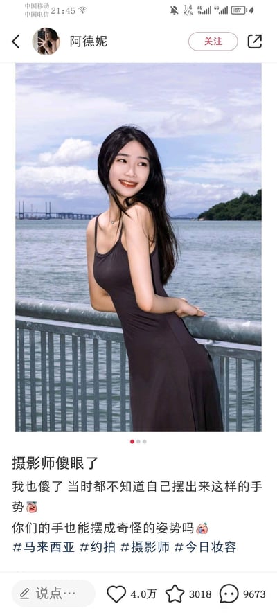 图片展示了一位身着黑色吊带长裙的女性站在海边栏杆旁的场景。她长发披肩，面带微笑，身体微侧倚靠在金属栏杆上，一只手撑着栏杆，背景可见平静的海面、远处的桥梁和绿树覆盖的小岛。图片下方配有文字 caption：“摄影师傻眼了 我也傻了 当时都不知道自己摆出来这样的手势😳 你们的手也能摆成奇怪的姿势吗🤔 #马来西亚 #约拍 #摄影师 #今日妆容”，表达了对拍照时无意间摆出奇怪手势的惊讶和调侃。