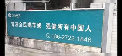 这是一张线下拍摄的隆力奇（Longrich）品牌宣传横幅照片，横幅为蓝绿色底配白色文字，核心宣传语为「普及全民喝羊奶 强健所有中国人」，下方标注了联系电话186-2722-1846。横幅设置在一处老旧建筑的窗下，背景可见斑驳的墙面与窗户，是品牌面向大众的接地气线下推广物料，图中内容无谐音梗、双关或暗示类表述。