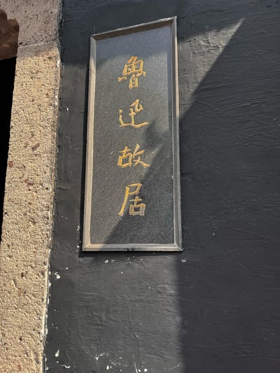 这是一张实拍的竖版牌匾照片，牌匾为深色磨砂底，搭配金色的竖排书法字体，写着「鲁迅故居」四个大字，安装在深色的墙面上。画面左侧是粗糙的石质门框，阳光照射下在牌匾和墙面上投下清晰的阴影，整体风格古朴庄重，是绍兴鲁迅故居的标志性标识牌。