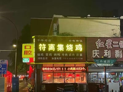 夜晚街道旁的一家餐馆外景，主要展示了一家名为'符离集烧鸡'的店铺招牌。招牌为黄色霓虹灯样式，十分醒目，上面写着'十年老店 口味保证'以及主营产品：烧鸡、酱鸡、烤鸭、牛肉、卤肉、凉菜等。下方还有特价信息：'特价24元/斤!!! 小鸡腿10元3个'。店铺旁边还有其他商家的招牌，包括'红星二锅'和'庆利副'，以及一个路牌显示'丹城路'。整个场景呈现出城市夜晚的烟火气。