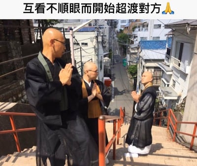 图片中三位光头男子身着黑色传统僧袍，其中一人外搭绿色袈裟，一人内搭黄色袈裟，第三人着纯黑僧袍配白色下装，三人站在红色栏杆的石阶上，身处狭窄的日式民居街道中（背景有密集的房屋、太阳能板和电线）。三人皆双手合十，呈祈祷姿势，却两两相对，表情严肃专注。图片顶部配有文字“互看不顺眼而开始超度对方🙏”，通过僧人本应慈悲的祈祷动作与“超度对方”的戏谑文字形成反差萌，营造出搞笑效果。