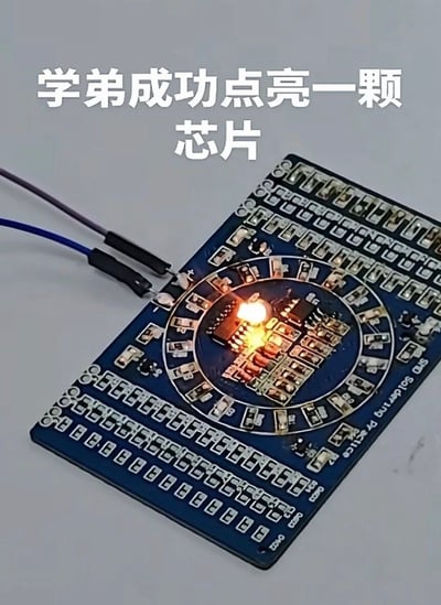 图片展示了一块蓝色电路板，上面布满密集的电子元件和线路。电路板中央有一个元件正在发光（或产生火花），疑似因短路或过载导致。上方配有文字“学弟成功点亮一颗芯片”，以幽默调侃的方式呈现“点亮”可能实际是损坏芯片的场景，带有校园实验室趣事的搞笑效果。
