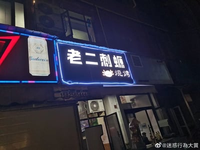 这是一张夜晚拍摄的街边烧烤店实拍照片，店铺的发光招牌十分醒目，主名称为“老二刺螈”，下方配有螃蟹图标和繁体的“燒烤”字样，其中“刺螈”是谐音“刺猿”，对应程序员的调侃称呼“程序猿”，是趣味十足的谐音梗用法。招牌左侧是带有Budweiser（百威）标识的广告灯牌，招牌下方标注了店铺联系电话15825654440。店铺门内可以看到有真实人物存在，整体场景是普通的街边餐饮店铺夜景。