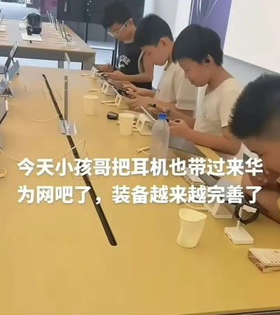 这张图片展示了几位小孩坐在电子产品体验店（类似苹果店）的展示桌前，聚精会神地用体验机进行娱乐，还自带耳机和饮料杯，仿佛在网吧包场一样。配文调侃说“今天小孩哥把耳机也带过来，真为网吧了，装备越来越完善了”，反映了一种利用免费科技体验店场地“变相”享受上网娱乐的幽默社会现象。

画面带有轻松的幽默意味，有“体验店变网吧”的现实双关，反映数字时代青少年的日常。